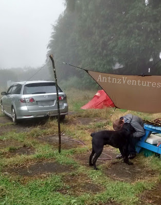 Antnz giving ADNZ Ben a morning cuddle, Antnz is sitting on a picnic table seat in right foreground. In the background is a car on left, a tent and shelter on right. It is a foggy morning.
