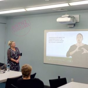 Annette Hansen standing to the left of a paused image of a NZSL video about Deaf Communications