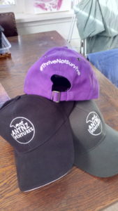 3 caps stacked. 2 bottom caps are black and green facing forward showing the AntnzVentures logo. Top purple caps shows the back with #ThriveNotSurvive