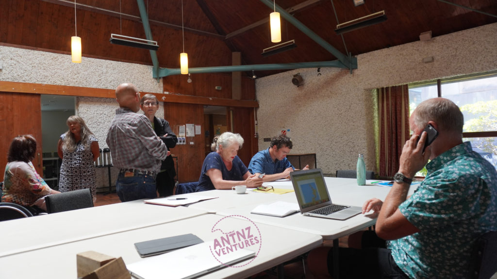 On the left of frame, Gerri Pomeroy in a wheelchair, Virginia Wilton is talking to Gerri.  Mark Benjamin and Anne Wilkson are standing talking to the right of Virginia. Marg Matheson and Shane McInroe are seated checking their phones, they are seated at a large table to the right of Mark and Anne.  To the right of the image John Taylor is seated, making a phone call and checking email on a laptop.  The background is a stucco hall.