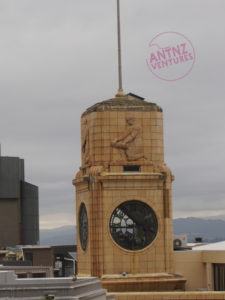 A faded limestone clock-tower.  The tower has a glass clock face, above the clock face there is a carved figure of a person kneeling on one knee with a bushel. 