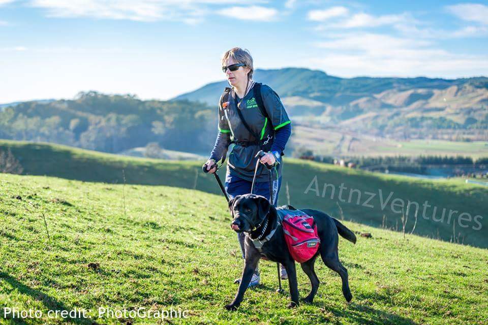 Antnz using walking poles, with ADNZ Ben licking his face. In the background are hills of Hawkes Bay