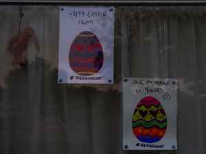 A picture of a window pane. On the top left is Kaha (Beanie Babie Kiwi) is handing from the top of the net curtail. Next to Kaha a A4 portrait page, with a colored Easter on it, this page has "Happy Easter From" written on the top and #nzegghunt on the bottom. To the right and lower than the 1st poster is a second poster of the same size. It also has a painted Easter egg (different colors than the first). On the top of the poster is written "Our Bubble To Yours", on the bottom "#nzegghunt" is also written.