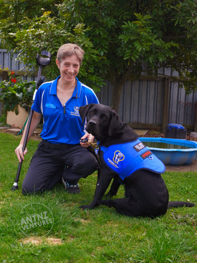 Antnz is crouched on the lawn to the left of the picture. She is holding a crutches in her right hand. To Antnz right, ADNZ Raven sits. They are both looking at the camera, Antnz has a cheesy grin on her face. Antnz is wearing an ADNZ blue tee shirt, ADNZ Raven has on her ADNZ blue Jacket.