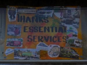 A hand made A2 poster. The background is painted, blended yellow, red and blue in different parts of the background. In the middle, in black lettering read "Thanks Essential Workers". Surrounding the lettering is a collage of different pictures of services and workers.