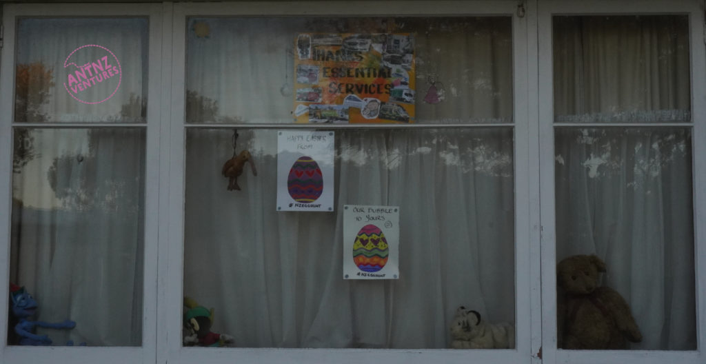 A picture of a white window, with curtains shut. Behind the glass are several teddy bears on the window sill, a stuffed Kiwi hanging from the frame, two colored-in Easter eggs and one Poster with text saying "Thanks essential workers" surrounded by smaller pictures representing different essential services