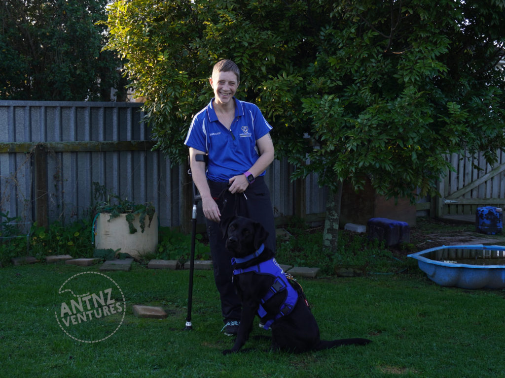 A photo of Antnz standing with a elbow crutch in her right hand. ADNZ Raven is sitting in front of Antnz. Taken in the back yard. Antnz is wearing a blue Assistance Dogs NZ polo shirt and Raven has her "work gear" on.