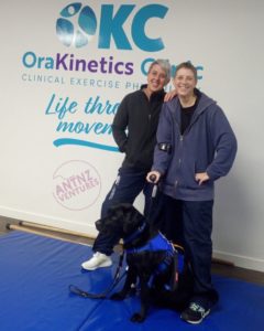 Antnz (right) and Tasha standing in front of an OraKinetics logo. ADNZ Raven is sitting in front of them looking off camera.