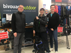 Antnz with 2 staff members from Bivouac Outdoors, Palmerston North in front of the counter. Antnz is holding a waterproof phone case. Antnz' elbow crutches can be seen to the side of them. ADNZ Raven is sitting in front of Antnz