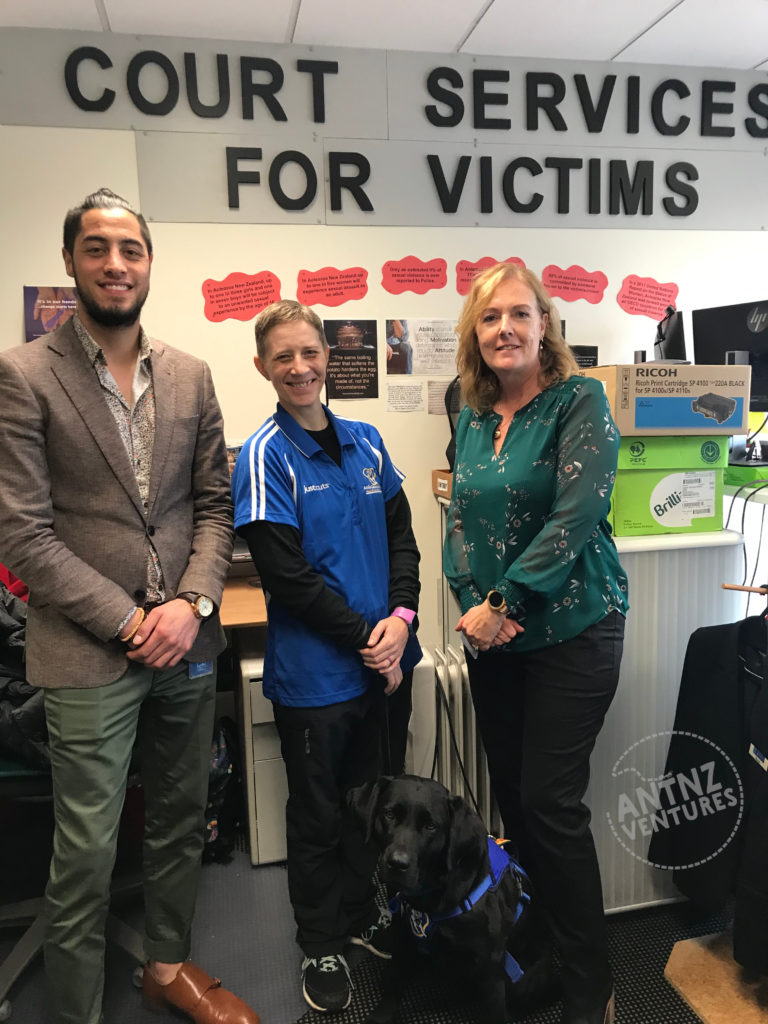 Antns is standing between Jamie (left) and Rose (right) in front of a Court Services for Victims sign. ADNZ Raven us sitting between Antnz and Rose.