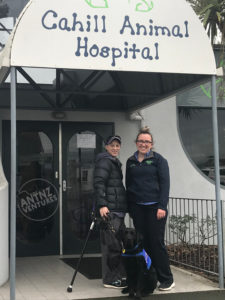 Antnz (on left) and Emily (right) from Cahill Animal Hospital standing in front of the clinic with the business logo over the enterance. ADNZ Raven is sitting in front of them.