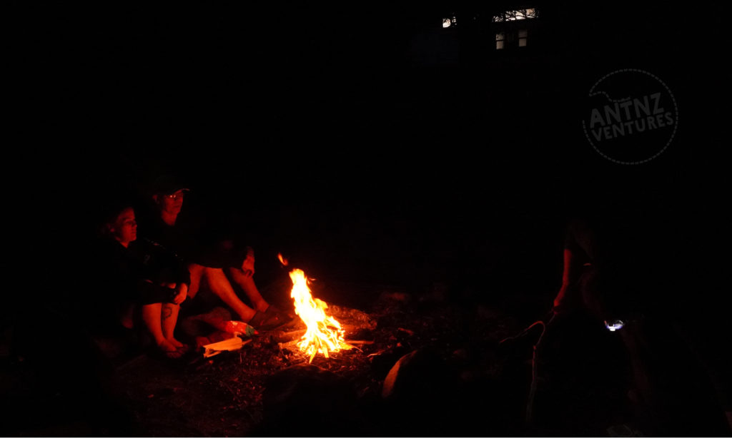 A picture taken at night of a campfire. The campground is in the foreground center of the picture. Natt and Peter sit to the left of the fire, to the right of the fire you can see a torch. In the the background are the lights at the hut.