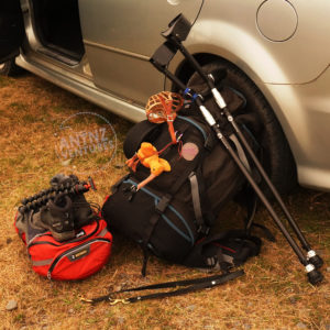 A large black backpack leans on the back wheel of a car. The front passenger door is open on the top, left of screen. To the left of the back are red, dog backpacks, with boots and a mini tripod left on stop. On the right of the backpack, a set of elbow crutches leans on the backpack. In front of the back pack, a black leather leash lies on the ground.