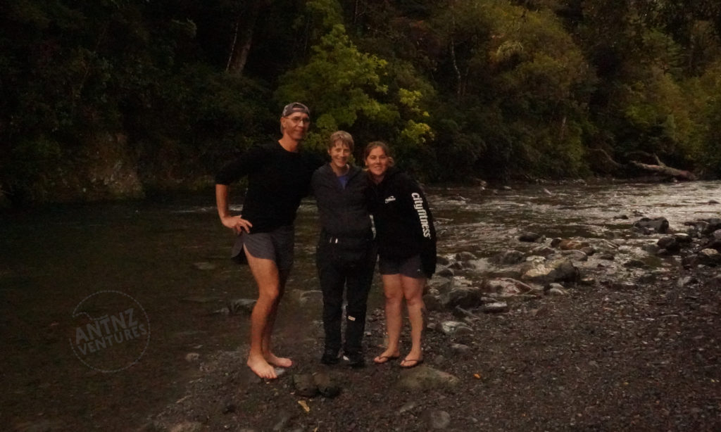 Picture Description: A picture of 3 people with arms around each other smiling at camera. They are standing by a river with native forest behind. From left to right stands Peter, Antnz and Natt.