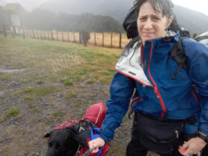 A selfie of Antnz and Raven. Antnz is wearing a blue rain jacket and has wet hair. ADNZ Raven is in the bottom of frame wearing a red rain jacket and saddle bags. Antnz has a slight grimace on her face.