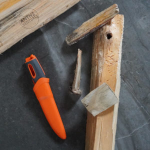 A bright orange knife is on the left of the frame in a sheath. To the right is a piece of hand width wood and 3 smaller pieces of wood shaped in wedges. On the piece of wood you can see marks of where it has struck the spine of the knife. The 3 wedges all have "mushrooming" on each end.