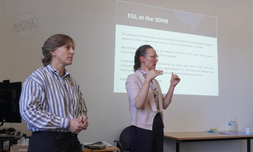 A photo of Antnz on the left, she is looking thoughtful. Carey-Ann is to the right of Antnz and is motioning with her hands to indicate levels. Behind and above them is a projected image of text. The top of the projected text reads "EGL at the 3DHB", the rest of the text is blurred