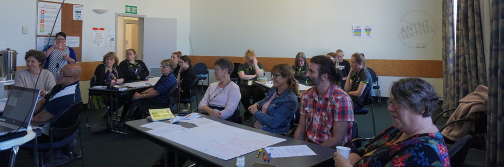 A group of people surround several tables in a classroom system. People in general are looking towards the left of the picture which is the front of the room.