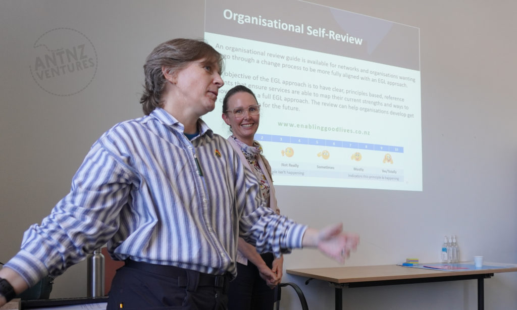 Antnz is slightly left of center, and is moving her hands. Behind her is Carey-Ann smiling. Behind them is a projector image of text. The top of the slide reads "Organisational Self-Review"