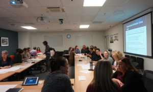 A room of people gathered in groups around a "board table" type set up. On one wall is a Projected image from a presentation. The title of the slide is "Organisational Self-Review