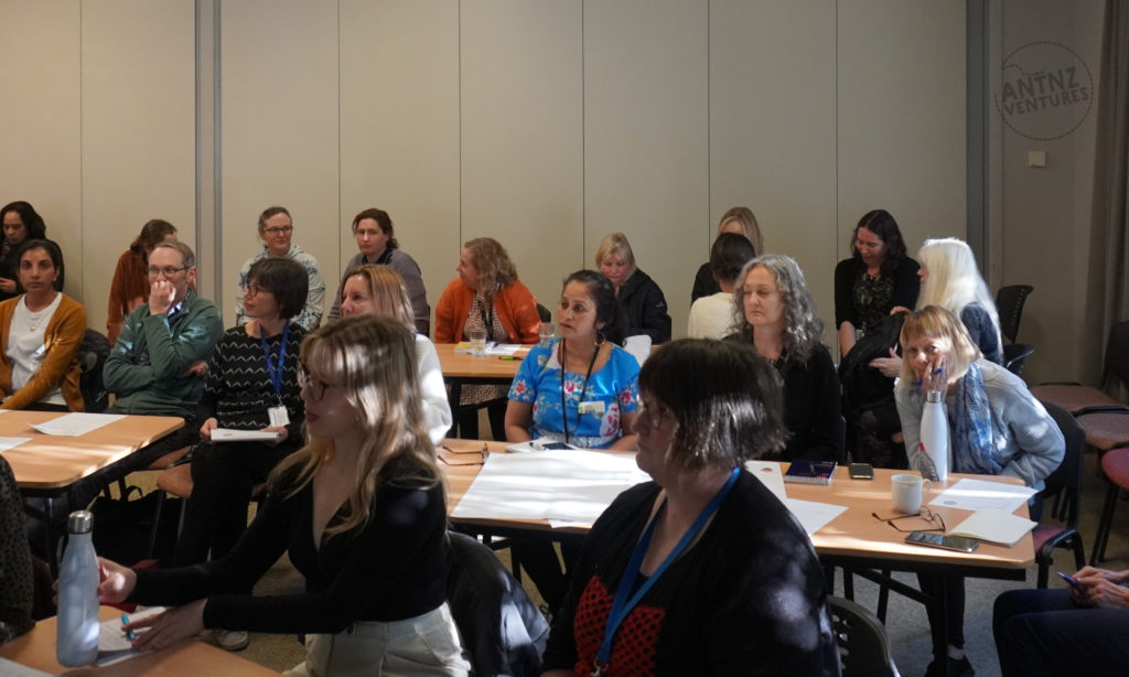 A group of people sitting at tables with paper and pens. Most people are looking in the same direction listening to the presenter.
