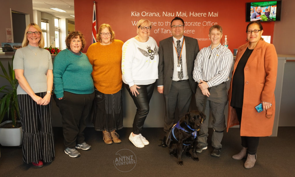 A picture of a group of people standing in a red room in a line facing the camera. The people from left to right are: Donna (Meeting Assistant), Rachel K (People First Rep), Karen (Family/Whanau Rep), Ally (Family/Whanau Core Group member), Tangi (Labour MP), Antnz (Disabled People's Rep), Lovey (Pasifika Rep). ADNZ Raven is sitting in front of Tangi & Antnz