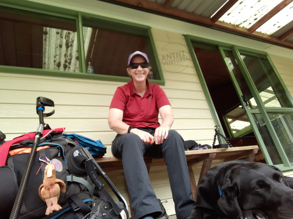 Antnz is sitting on a bench seat outside a building, the picture is taken from ground level looking up. To the left of Antnz is a backpack,a orthotic on a boot and an elbow crutch. To the right of Antnz, in the foreground lying on the ground is ADNZ Raven, a black Labrador