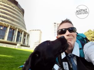 In the background left of the picture is the side of NZs Beehive. To the right is Antnz from shoulders up. I have light complexion, with short spiky light hair, wearing sunglasses, with a screwed up face. Between the Beehive & Antnz is ADNZ Raven, a black Labrador, licking Antnz face