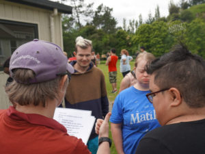 On the left of the picture is Antnz from behind, wearing a purple cap, with #ThriveNot Survive embroidered on it. Antnz is giving directions from a sheet she is holding. Next to Antnz, on the right of frame is the side of a person's head, in the background are some people listening to Antnz, further in the background more people standing around can be seen.
