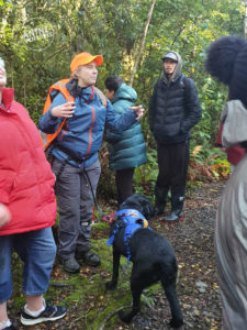 A NZ bush background. Antnz is standing wearing a blue jacket, a orange high-visibility cap and over her pack; she is gesturing as she explains something to a group of people.. Antnz is also wearing a waist leash attached to ADNZ Raven, who is looing away from the camera