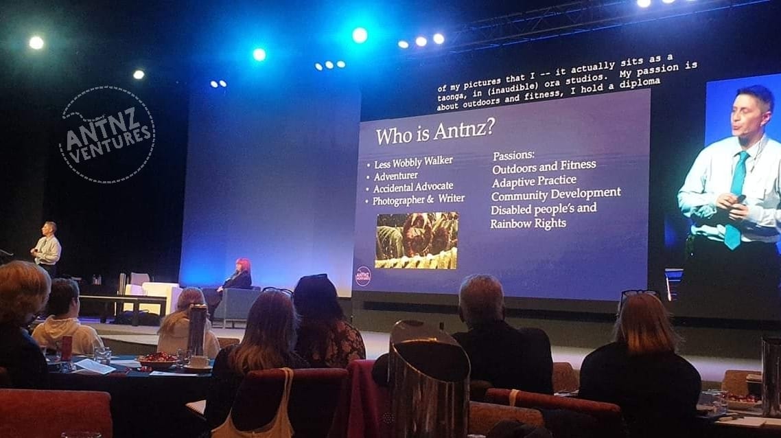 A wide angle shot of a stage from a table. On the left is a lecturn that Antnz is standing at. To the right of the lecturn on the stage are 2 chairs with a coffe table in front of the chairs with a pink haired woman in the right hand chair. A black lump, which is ADNZ Raven is between the coffee table and chairs. To the right, taking up most of the background is a screen, the majority containing a PowerPoint. The right hand third of the screen is an expressive picture of Antnz wearing a white shirt with blue pinstripes and a tie.