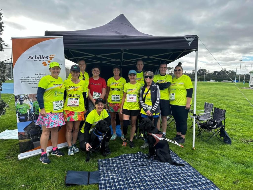 A group of people wearing bright yellow Achilles t-shirts, some with the word "Guide" on them. Antnz is crouching at the front with ADNZ Raven. Guide Dog Devon sits next to Raven. One woman in the front row has a below the knee prosthetic