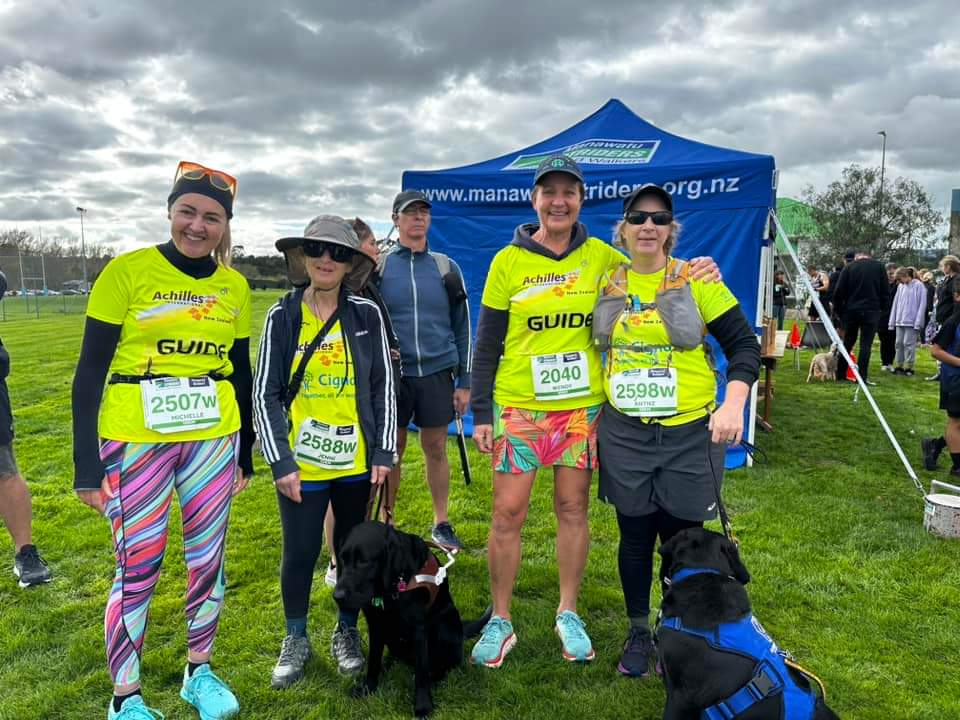 4 women in bright yellow stand in pair with 2 black Labradors