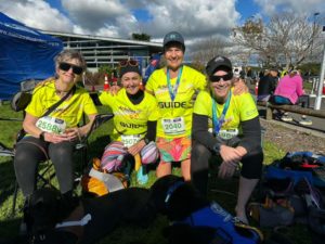 Jenny on the left and Antnz on the right are sitting on seats, between them crouching is Shell and Wendy our Guides. Everyone is smiling. Antnz and Wendy are wearing medals. Guide Dog Devon and ADNZ Raven are lying in front of them.