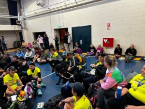 A group of people sitting in a sports hall, there are some people sitting in seats, and most of the floor. There is a clump of people in bright yellow Achilles t-shirts smiling, there are 2 black dogs sitting in the middle of the yellow t-shirts.