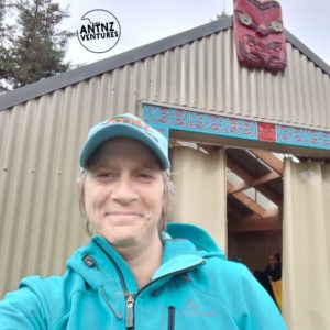 A selfie of Antnz standing in front of a corrugated iron whare (shelter). Antnz is smiling at the camera and wearing NATO blue cap with orange writing and a teal jscket. Tgr door frame of tge whare has blue and red carvings, with a carved figurehead above the door at the apex of the roof
