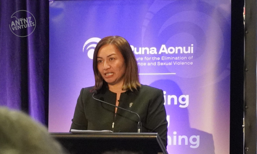 Minister Marama Davidson standing behind a lectern, in front of a purple Te Puna Aonui banner. She is speaking