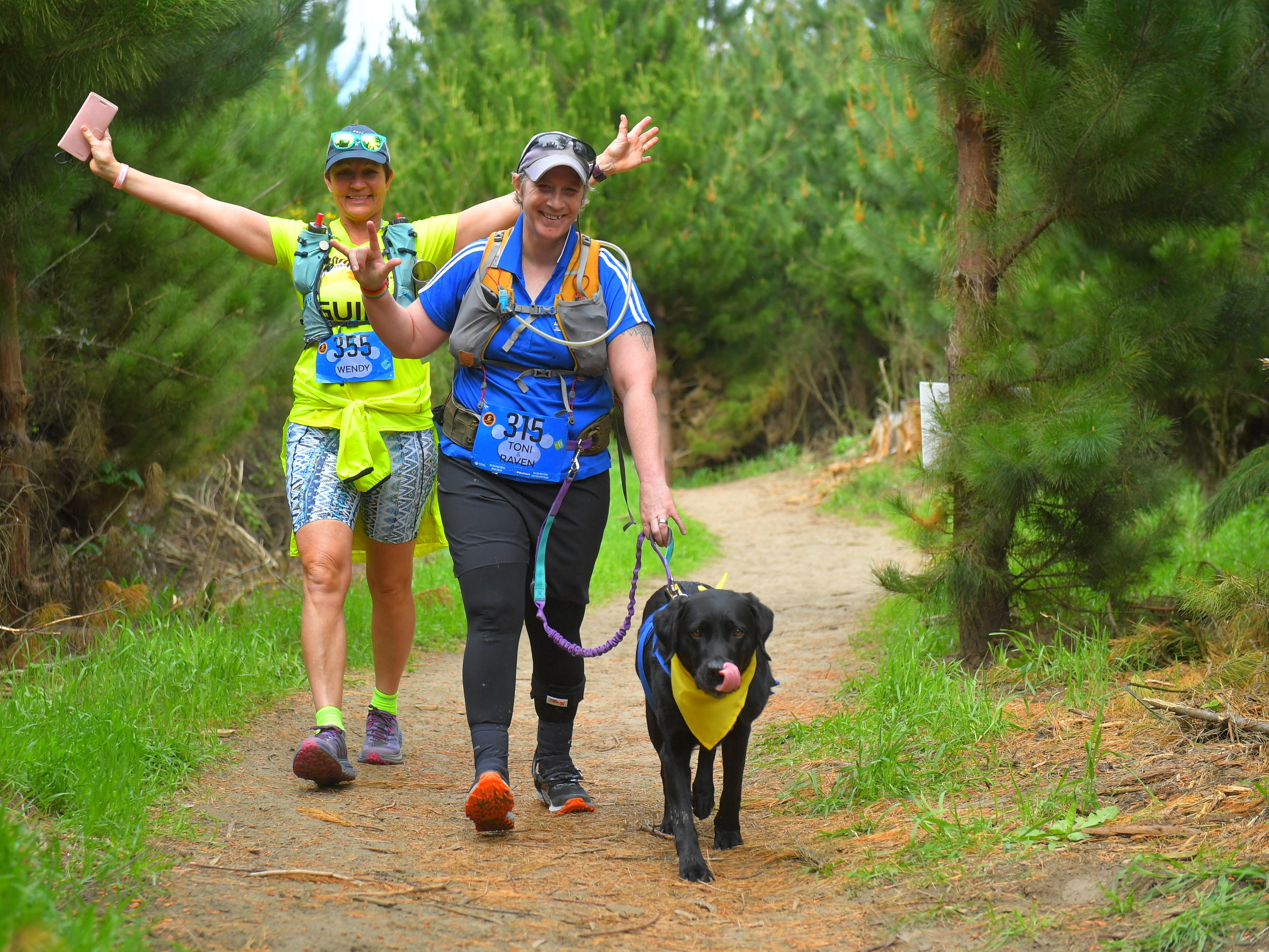 Antnz, ADNZ Raven and Wendy walking on a track with grass edges. Antnz is smiling, doing the international sign language for "I love you", wearing a blue shirt and is smiling. Wendy is wearing Achilles yellow and has her arms up in celebration and smiling. Raven is wearing blue jacket and yellow bandana and has her tongue out licking her lips