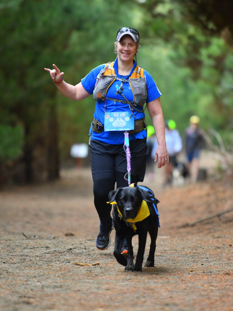 Antnz and Raven are walking towards the camera. Antnz is smiling and doing the international sign language for "I love you". She is wearing a blue t-shirt, black tights, yellow running vest and purple cap with sunglasses resting on top. Raven is concentrating, on a long lead with her ears bobbing slightly with her movement.