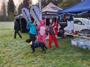 Antnz, ADNZ Raven and 2 members of the FurtherFaster team. Antnz is wearing a jacket and her running tights and is doing the international sign language of I Love You. Jules, to the right of Antnz is wearing a Pink Panther Onsie pulling a thumbs up and holding a coffee cup. To the right of Jules is Hanna (?) wearing a Santa onsies and pulling thumbs up!
