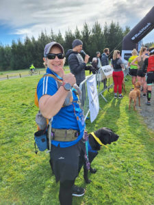 A picture of Antnz and ADNZ Raven. Antnz is wearing running tights and shorts, a blue t-shit, purple cap and a wide belt. Raven is wearing her blue working harness and a yellow bandana. Antnz is smiling and pulling a thumbs up at the camera. In the background is a man wearing a beanie and puffy jacket, taking in to a microphone. To the right of him are several people and a dog.