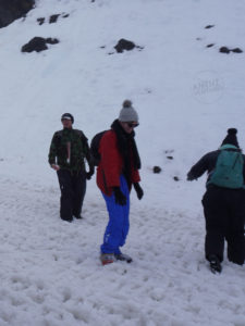 A young man wearing blue snow pants, a red jacket, black scarf, sunglasses, black gloves and a grey beanie with a pompom. In the back ground is another man wearing an army jacket, cap with black beanie, glasses and black snow pants, he has a snowball in hand and a white cane folded and attached to his jacket