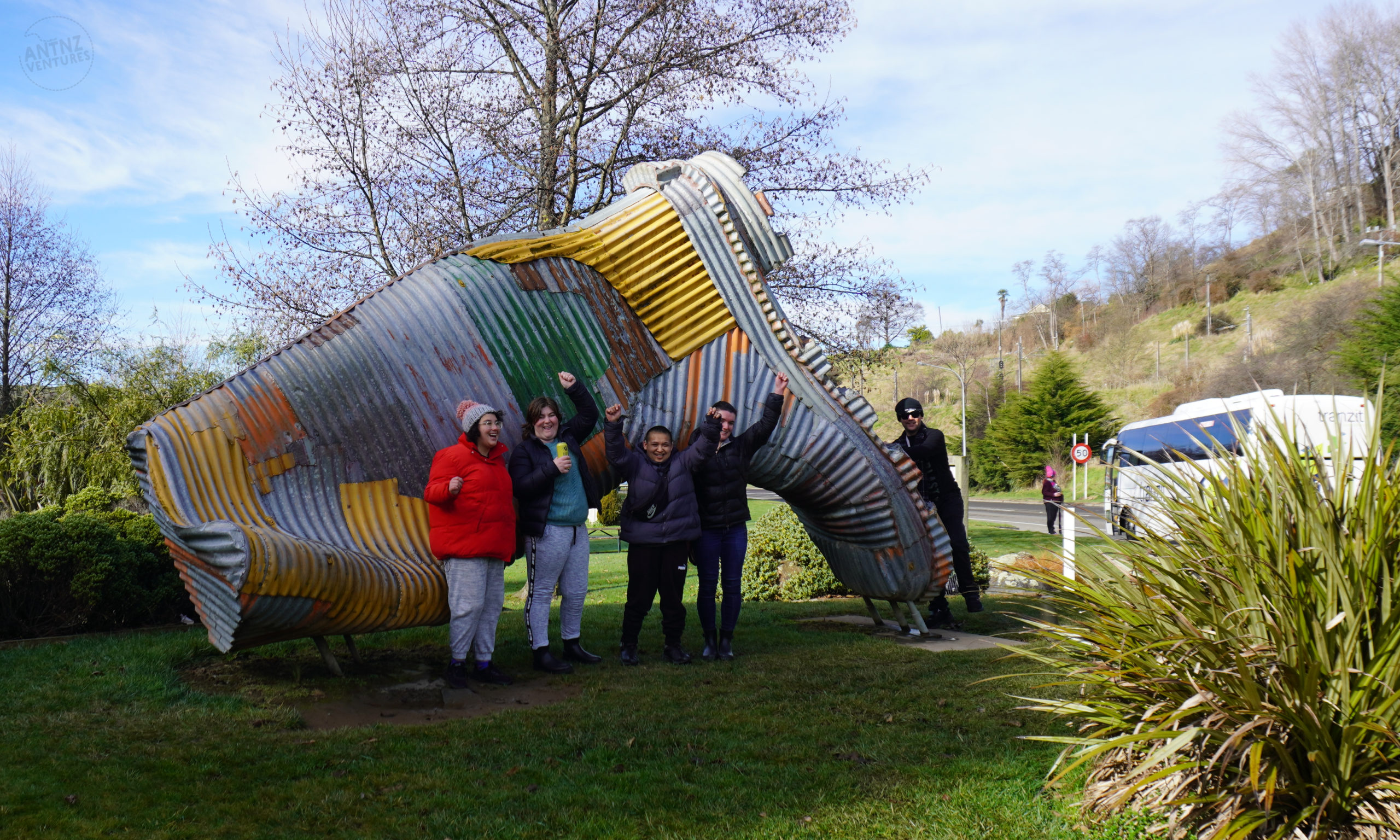 A group of 4 people wearing winter clothing standing in front of a large, corrugated iron gumboot lying with its toes towards the ground. At the toes of the boot is another person wearing winter clothing leaning on the boot. All the people look happy ans some are raising their arms in celebration