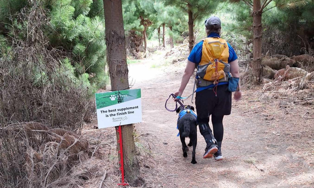 Antnz and Raven walking away from camera, down a slight hill in a pine forest. The is a sign to Antnz left that reads "The best supplement is the finish line.