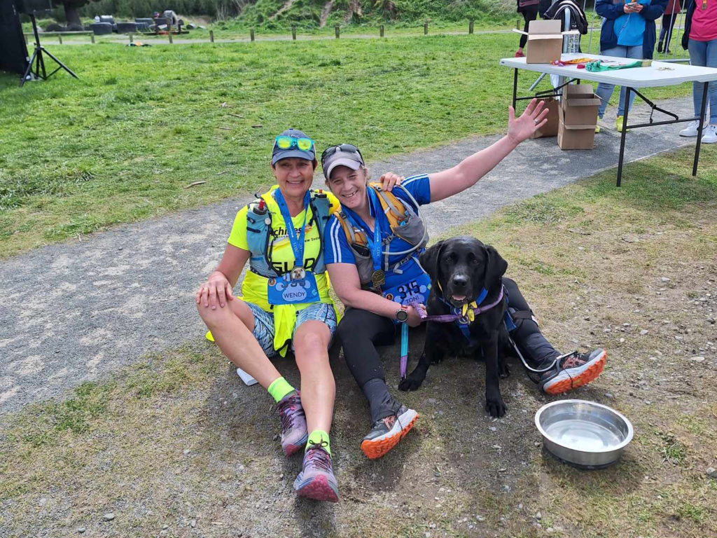 Wendy on the right in Achilles yellow with her sunglasses on the cap on her head, she is wear a medal on a blue ribbon around her neck and has one arm around Antnz. Antnz is smiling with her arm up in celebration, also wearing a medal on a blue ribbon. Raven is sitting looking at the camera. There is a water bowl on the ground next to Antnz' feet.