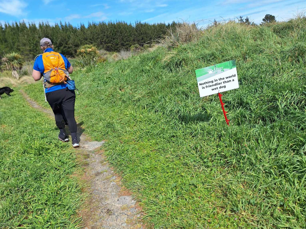 Antnz walking away from camera on narrow path in grass. To the right of Antnz is a sign that reads "Nothing in the world is friendlier than a wet dog"